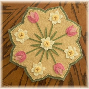 May include: A beige felt circle with a scalloped edge, featuring a central cluster of white daffodils with yellow centers and green leaves. Surrounding the center are six pink tulips and six white daffodils with yellow centers, all with green stems.