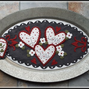 Peut inclure: Un dessous de plat en forme de cœur en feutre gris avec trois cœurs rouges cousus dessus. Les cœurs ont des centres blancs et sont brodés avec les mots "Je t'aime". Le dessous de plat est sur un plateau ovale en argent avec des poignées.