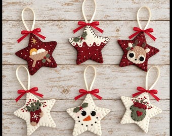 May include: Six star-shaped felt ornaments featuring holiday designs including a manger, a pudding, a reindeer, a stocking, a snowman, and a mitten. Each ornament is stitched with white thread, topped with a red ribbon bow, and a cream hanging loop.