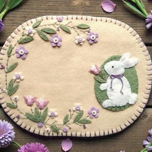 May include: Oval-shaped beige felt placemat with a stitched border. Features embroidered floral vines with purple flowers and green leaves. A white felt bunny holds a pink flower, set on a green oval.