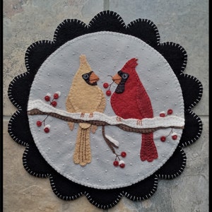 May include: A felt applique featuring two cardinals perched on a snow-covered branch with red berries. The birds are in shades of red and tan, set against a gray background with white dots. The design is framed by a black scalloped edge.