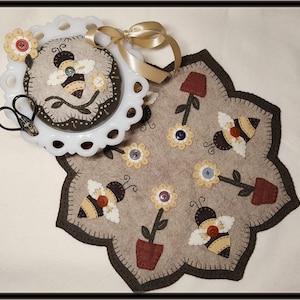 May include: A beige felt bee themed pincushion with a white lace trim and a brown felt bee with yellow stripes. The pincushion is on a white pedestal with a gold ribbon. The pincushion is surrounded by a beige felt bee themed needle minder with brown felt bees, yellow flowers, and brown pots.