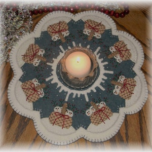 May include: A blue felt Christmas table runner with a white border and a snowflake design. Eight snowmen holding baskets are arranged around a clear glass candle holder with a lit candle.
