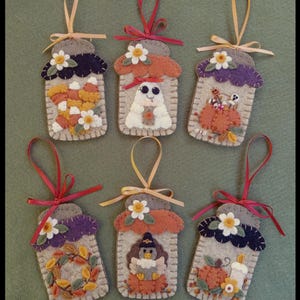 Könnte beinhalten: Sechs Filz-Ornamente in Gläserform mit verschiedenen Herbstmotiven. Jedes Ornament hat einen grauen Deckel, ein Band zum Aufhängen und ist mit Kürbissen, Geistern, Zuckermais und Blumen verziert. Die Ornamente sind in Orange-, Lila- und Cremetönen gehalten.