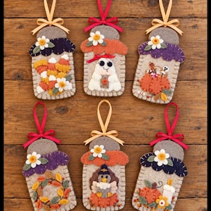 May include: Six felt ornaments shaped like mason jars, each featuring a different seasonal motif including candy corn, a ghost, pumpkins, an autumn wreath, a turkey, and a candle. Each ornament has a ribbon loop and a small felt flower accent.