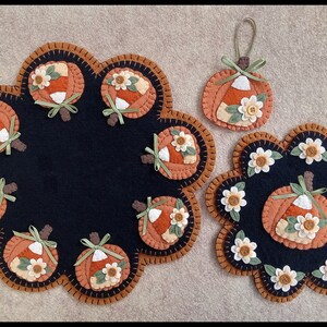 May include: Three felt pumpkin decorations with candy corn and flower accents. The largest decoration is a round table topper with a black background and eight pumpkins around the edge. The smaller decorations are round and have a black background with a single pumpkin in the centre.