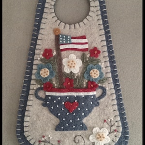 May include: A felt door hanger with a patriotic theme. It features a blue vase with white polka dots, a red heart, and an American flag. The vase is filled with red, white, and blue flowers. The hanger has a blue border and a hole at the top.