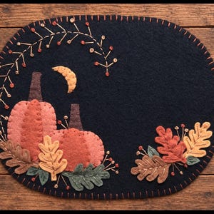 May include: An oval black felt mat featuring hand-stitched applique designs of two orange pumpkins with brown stems, a yellow crescent moon, and various autumn-colored oak leaves and berries arranged in a decorative seasonal pattern.