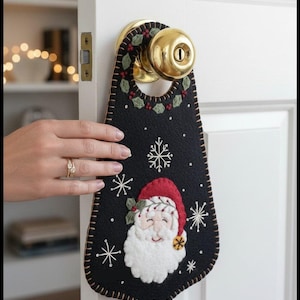 May include: A black felt door hanger featuring an embroidered Santa Claus face with a red hat, white beard, and green holly accents. The design includes white stitched snowflakes and a decorative border with tan thread, hanging from a brass doorknob.
