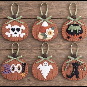May include: Six felt pumpkin ornaments with green ribbon hangers. Designs include a skull with heart eyes, candy corn with a flower, a bubbling cauldron, the word BOO with a star, a white ghost, and a black boot with decorative green sprigs.