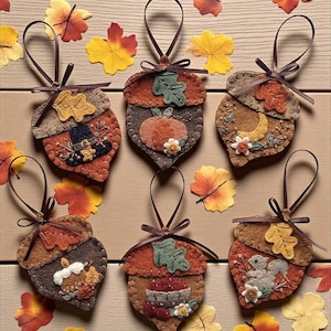 Può includere: Sei decorazioni a forma di ghianda in feltro con nastri marroni. Ogni decorazione presenta applicazioni a tema autunnale, tra cui foglie, zucche e uno scoiattolo. Le decorazioni sono nei toni del marrone, arancione e giallo, con cuciture decorative.