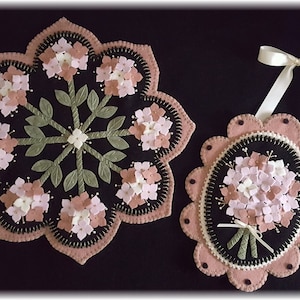 Puede incluir: Dos tapices de pared de fieltro con diseños de flores rosas y blancas. El más grande es un diseño redondo con un patrón de estrella y el más pequeño es un diseño ovalado con un solo ramo de flores.