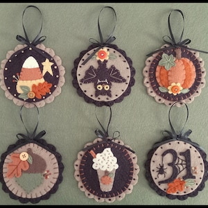 May include: Six felt Halloween ornaments with black ribbon hangers. The ornaments feature a candy corn, bat, pumpkin, acorn, milkshake, and a spider web with the number 31.