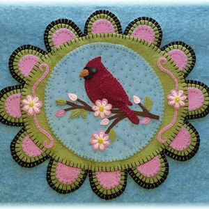 May include: A felt wall hanging with a red cardinal perched on a branch with pink flowers. The cardinal is surrounded by a green circle with a black and pink scalloped border.