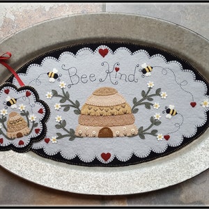 May include: A gray felt bee-themed doorstop with a beehive, bees, and flowers. The doorstop has the text "Bee Kind" on it. The doorstop is on a metal tray.