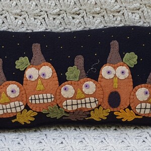 Puede incluir: Una almohada de fieltro negro con una fila de calabazas de fieltro naranja con caras divertidas. Las calabazas tienen hojas y tallos de fieltro verde. La almohada tiene un borde de fieltro negro con pompones negros.
