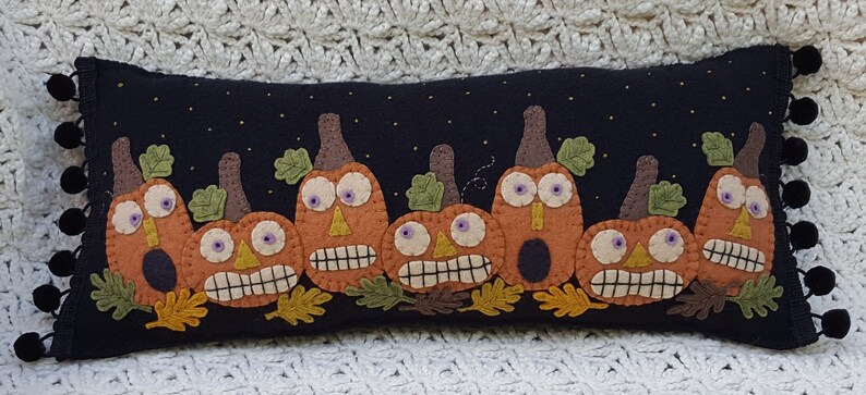 Puede incluir: Una almohada de fieltro negro con una fila de calabazas de fieltro naranja con caras divertidas. Las calabazas tienen hojas y tallos de fieltro verde. La almohada tiene un borde de fieltro negro con pompones negros.