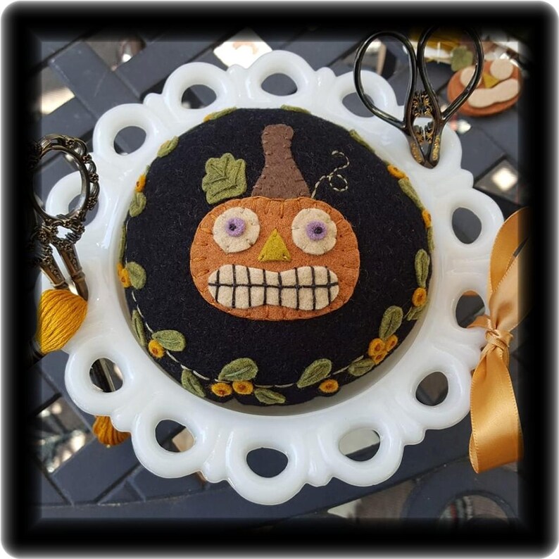 Puede incluir: Un coj&iacute;n de alfileres de cer&aacute;mica blanca con una parte superior de fieltro negro que presenta una calabaza naranja bordada con una sonrisa de dientes y ojos morados. El coj&iacute;n de alfileres est&aacute; en un plato blanco con un borde festoneado.