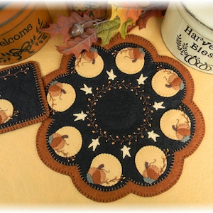 May include: A black felt table runner with a scalloped edge and a design of pumpkins and stars. The runner is surrounded by fall-themed decorations, including a wooden box with the word "Welcome" and a wooden box with the words "Harvest Blessings".