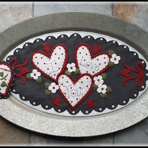 Puede incluir: Una decoración de fieltro con temática de San Valentín con tres corazones blancos con lunares que dicen "I Love You" sobre un óvalo gris oscuro. La decoración está sobre una bandeja de metal con un adorno en forma de corazón.