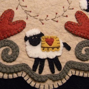 Könnte beinhalten: Eine Filz-Handarbeit mit einem weißen Schaf mit schwarzen Hufen und einem gelben und roten Sattel. Das Schaf ist umgeben von grauen Filz-Wirbeln und zwei roten Filz-Herzen.