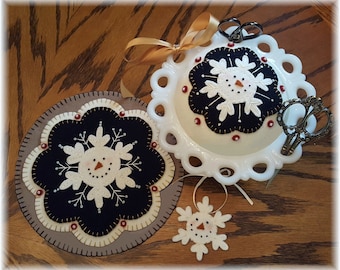 May include: Three felt snowflake ornaments with a snowman face. The ornaments are white with black stitching and red bead accents. The ornaments are on a wooden surface with a white ceramic dish and gold ribbon.