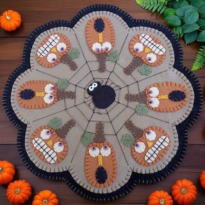 May include: A Halloween-themed felt placemat featuring a central spiderweb design with a black spider. The placemat has a gray background with orange pumpkin-shaped figures with various facial expressions. The border is black and scalloped.