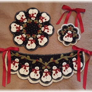 May include: A set of three felt snowman ornaments with red bows. The ornaments are in the shape of a circle, a half-circle, and a long rectangle. The ornaments are decorated with green holly leaves and red berries.