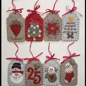May include: Eight handmade felt Christmas decorations with red ribbon hangers. Each tag-shaped ornament features a different festive design, including a heart, Christmas tree, poinsettia, and snowman. The ornaments are in shades of grey, red, and green.