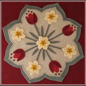 Peut inclure: Une suspension murale en feutre beige avec un motif en étoile. Le centre de l'étoile a une fleur jaune avec un centre jaune. L'étoile est entourée de six fleurs jaunes avec des centres jaunes et six tulipes rouges avec des centres jaunes.