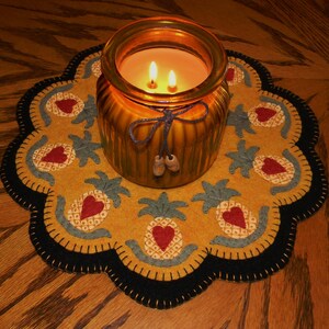 May include: A yellow and black felt pineapple-themed candle mat with a glass candle jar in the center. The candle jar has a brown lid and a brown string tied around the jar. The candle is lit and the flames are visible.