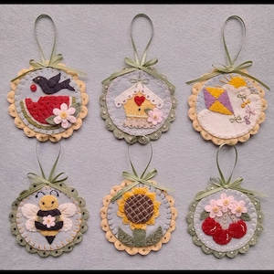May include: Six felt ornaments with different designs, including a watermelon, a birdhouse, a kite, a sunflower, a bee, and cherries. Each ornament is attached to a green ribbon and has a scalloped edge.