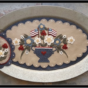 Peut inclure: Une suspension murale en feutre sur le thème patriotique. Elle présente un vase bleu et blanc avec des cœurs rouges, rempli de fleurs bleues et blanches et deux petits drapeaux américains. Le vase est entouré de feuilles vertes et de fleurs blanches. La suspension murale est sur un plateau en métal avec des poignées.