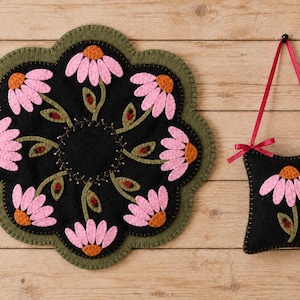 May include: A round, scalloped-edge felt mat and a small, square felt hanging ornament. Both items are black with pink and orange daisy-like flower appliqués, green stems, and small red ladybug accents, featuring visible blanket stitching.