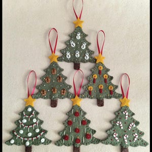 Könnte beinhalten: Sechs handgefertigte Weihnachtsbaum-Ornamente aus Filz. Jeder grüne Baum hat einen braunen Stamm, einen gelben Stern als Spitze und ein rotes Band zum Aufhängen. Die Bäume sind mit verschiedenen festlichen Designs verziert, darunter Schneemänner, Lebkuchenmänner, Kerzen, rote Kugeln und Zuckerstangen.