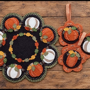 May include: A large black felt mat with a scalloped edge, decorated with orange and white felt pumpkins and a central leaf wreath, alongside three smaller matching felt pumpkin ornaments, each featuring a checkered fabric hanging loop.