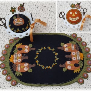 May include: A black felt needlepoint runner with a green border and a wreath of green leaves in the center.  The runner is decorated with five orange felt pumpkins with stitched-on eyes and mouths.