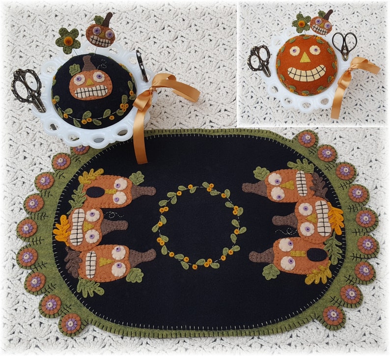 Puede incluir: Un camino de mesa de fieltro negro con un borde verde y una corona de hojas verdes en el centro. El camino de mesa est&aacute; decorado con cinco calabazas de fieltro naranja con ojos y bocas cosidos.