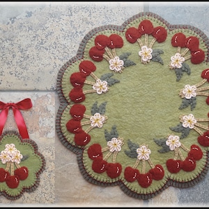 May include: A green felt rug with a cherry and flower design. The rug is round and has a scalloped edge. A smaller version of the rug is also shown, hanging from a red ribbon.