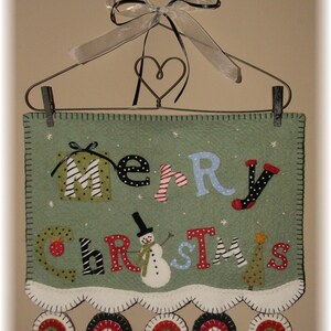 Op de afbeelding: Een groene vilt kerstbanner met de woorden "Merry Christmas" in kleurrijke letters. In het midden van de banner staat een sneeuwpop. De banner hangt aan een draadhangertje met een hartvormige draadlus. Er hangen acht ronde viltversieringen aan de onderkant van de banner.