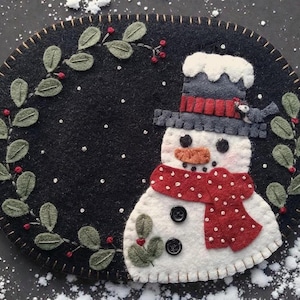 Puede incluir: Un aplique circular de fieltro negro con un muñeco de nieve blanco que lleva un sombrero de copa gris oscuro con una banda roja y una bufanda roja de lunares. El muñeco de nieve tiene ojos de botón negros, una nariz naranja y dos botones negros. Una corona de hojas verdes con bayas rojas rodea al muñeco de nieve.