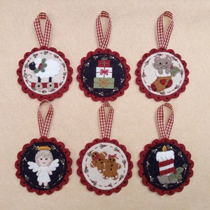 May include: Six felt Christmas ornaments with red and white gingham ribbon hangers. The ornaments feature a sheep pulling a wagon, a stack of presents, a cat in a stocking, a gingerbread couple, an angel, and a candle.