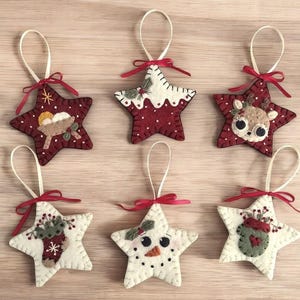 Puede incluir: Seis adornos de estrella de fieltro con diferentes temas navideños. Los adornos son rojos, blancos y verdes con detalles bordados. Los temas incluyen un muñeco de nieve, un reno, una media, una escena de la natividad y una casa de jengibre.