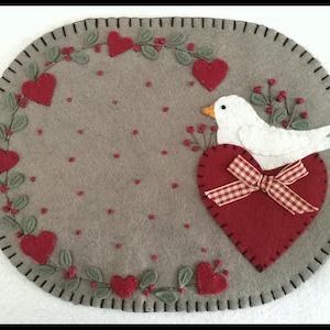 May include: A gray felt placemat with a stitched border, featuring red hearts, green leaves, and a white bird perched on a red heart with a plaid bow. The design includes red berry accents.