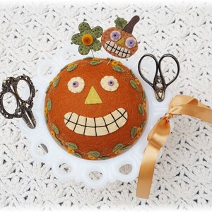 Puede incluir: Un coj&iacute;n de alfileres de cer&aacute;mica blanca con forma de calabaza con una cara sonriente, ojos morados y una nariz amarilla. El coj&iacute;n de alfileres est&aacute; decorado con hojas verdes y flores naranjas. Hay dos pares de tijeras y una cinta marr&oacute;n en el coj&iacute;n de alfileres.