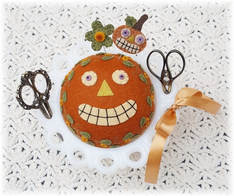 Puede incluir: Un coj&iacute;n de alfileres de cer&aacute;mica blanca con forma de calabaza con una cara sonriente, ojos morados y una nariz amarilla. El coj&iacute;n de alfileres est&aacute; decorado con hojas verdes y flores naranjas. Hay dos pares de tijeras y una cinta marr&oacute;n en el coj&iacute;n de alfileres.