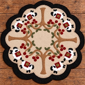 May include: A round felt mat with a scalloped edge featuring a beige center, a black border, and a repeating pattern of brown tree branches, green leaves, red berries, and white sheep with black heads and red blankets on their backs.