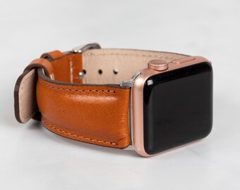 May include: A rose gold Apple Watch with a brown leather strap. The strap is made of stitched leather and has a silver buckle. The watch face is black. The watch is displayed on a white surface.