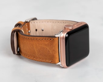 May include: A brown leather watch strap attached to a rose gold Apple Watch. The watch strap is made of distressed leather and has a silver buckle. The watch face is black.