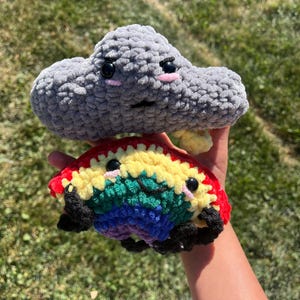 May include: A gray crocheted cloud with a frowning face is holding a rainbow crocheted cloud with a smiling face. Both clouds have black eyes and pink cheeks.